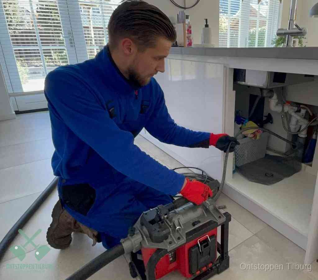 Een monteur met rode handschoenen knielt in een keuken en voert een elektrische ontstoppingsveer in tijdens het professioneel afvoer ontstoppen in Tilburg